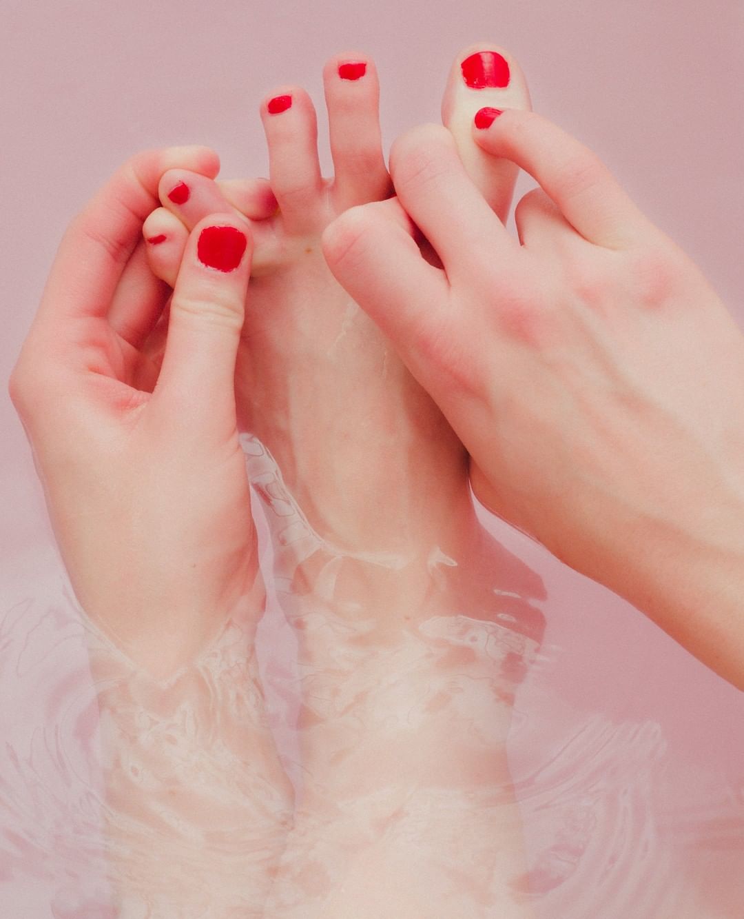 Close-up of female hands gently touching toes with red nail polish, feet submerged in water. Beauty shot for foot care or self-care concept.