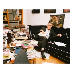 An elderly man seated on a dark velvet sofa surrounded by tall stacks of books in a living room interior.