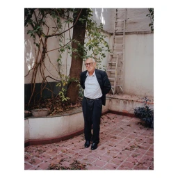 Portrait of an older man wearing glasses and a dark suit jacket standing in an outdoor courtyard with fallen leaves