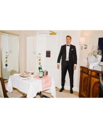 Man in a black tuxedo standing in an interior hotel room next to a table set with white cloth and food.