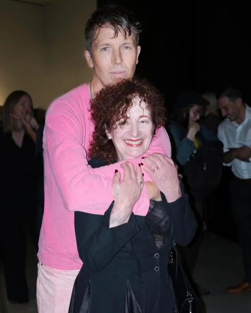 Portrait of a blonde adult male hugging an adult female with curly red hair while attending a social event. Attendees are blurred in the background.