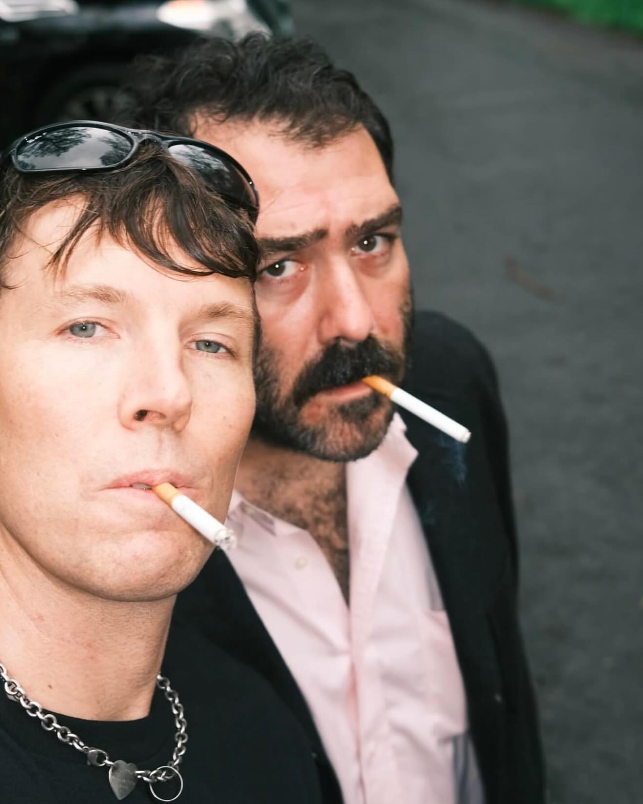 Two men smoking cigarettes outdoors; the foreground subject wears sunglasses on his head and a heart pendant necklace.