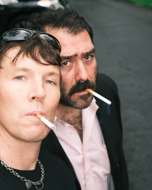 Two men smoking cigarettes outdoors; the foreground subject wears sunglasses on his head and a heart pendant necklace.