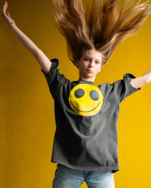 Young woman with long hair jumps in front of a yellow background, wearing a T-shirt with a yellow smiley face graphic.