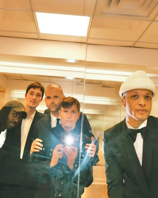 Group of five adult men in formal attire posing indoors, reflected in a mirror, creating a studio-style shot with a focus on fashion and entertainment.