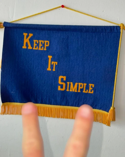 Blue banner hanging indoors, featuring the text 'Keep It Simple' in yellow lettering, adorned with yellow fringe and a partial view of a hand.