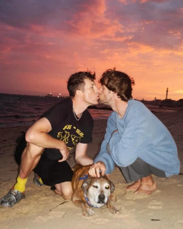 Two men kneeling and kissing on a sandy beach at sunset, with a brindle dog sitting between them.