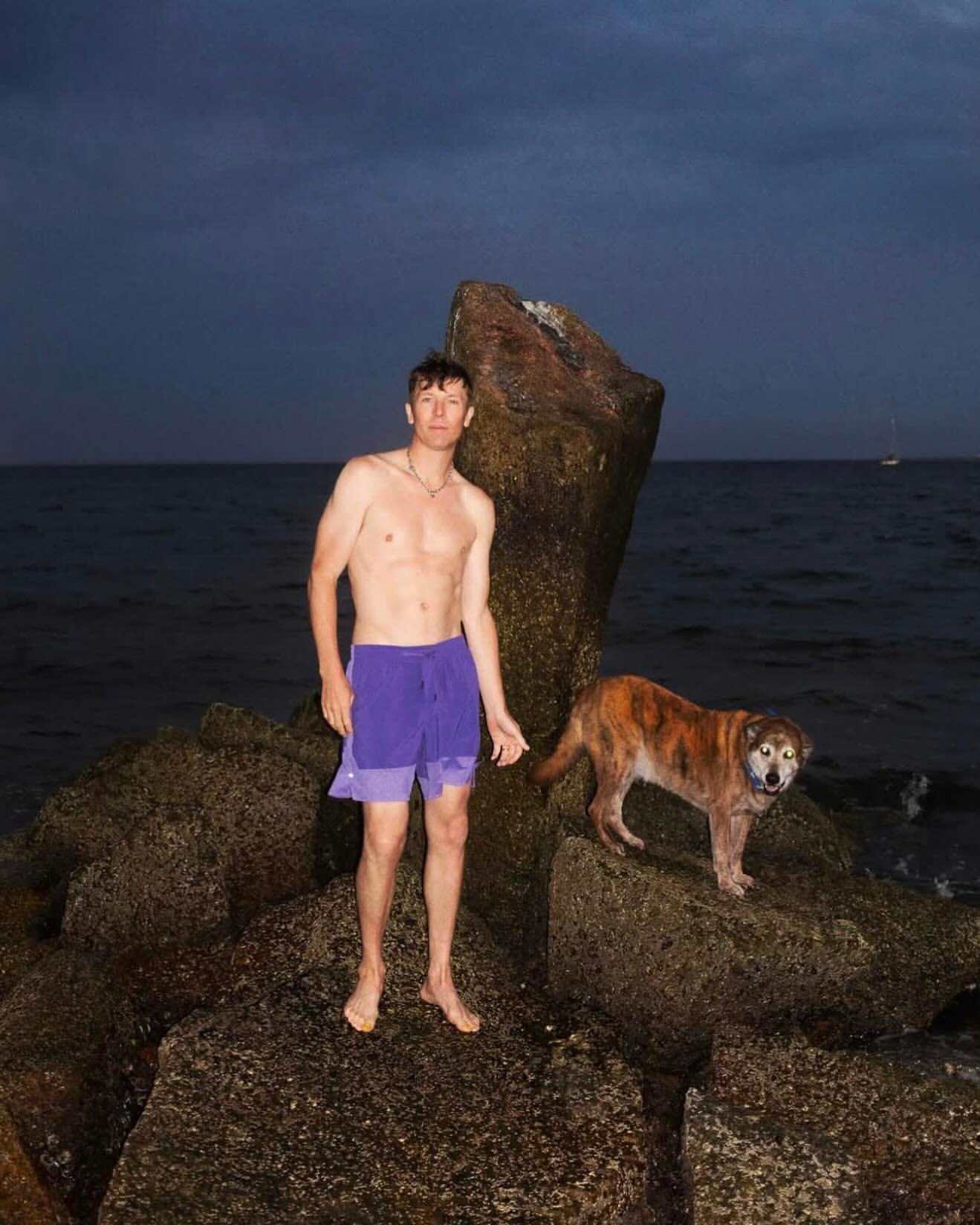A shirtless man in swim trunks stands on a rocky beach with a dog at dusk. The ocean and sky are visible in the background.