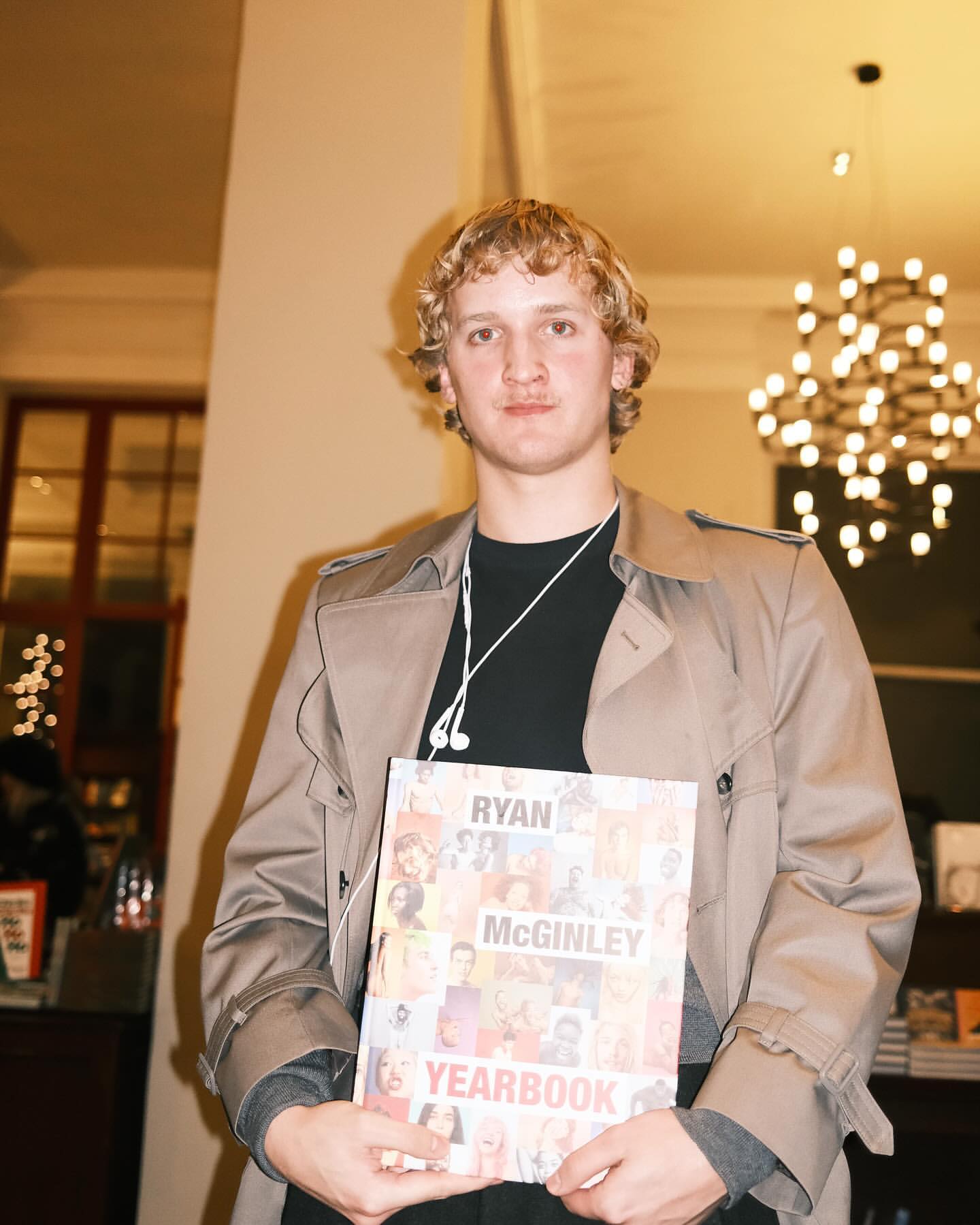 Young man in trench coat holds Ryan McGinley's Yearbook in an indoor setting, chandelier visible in background, close-up shot.