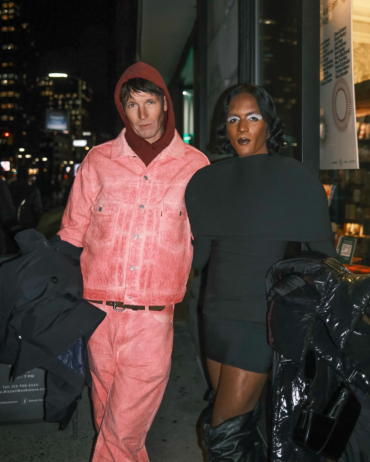 Two people pose in an urban street setting at night. One wears a pink denim suit, and the other has on a black dress with dramatic sleeves.