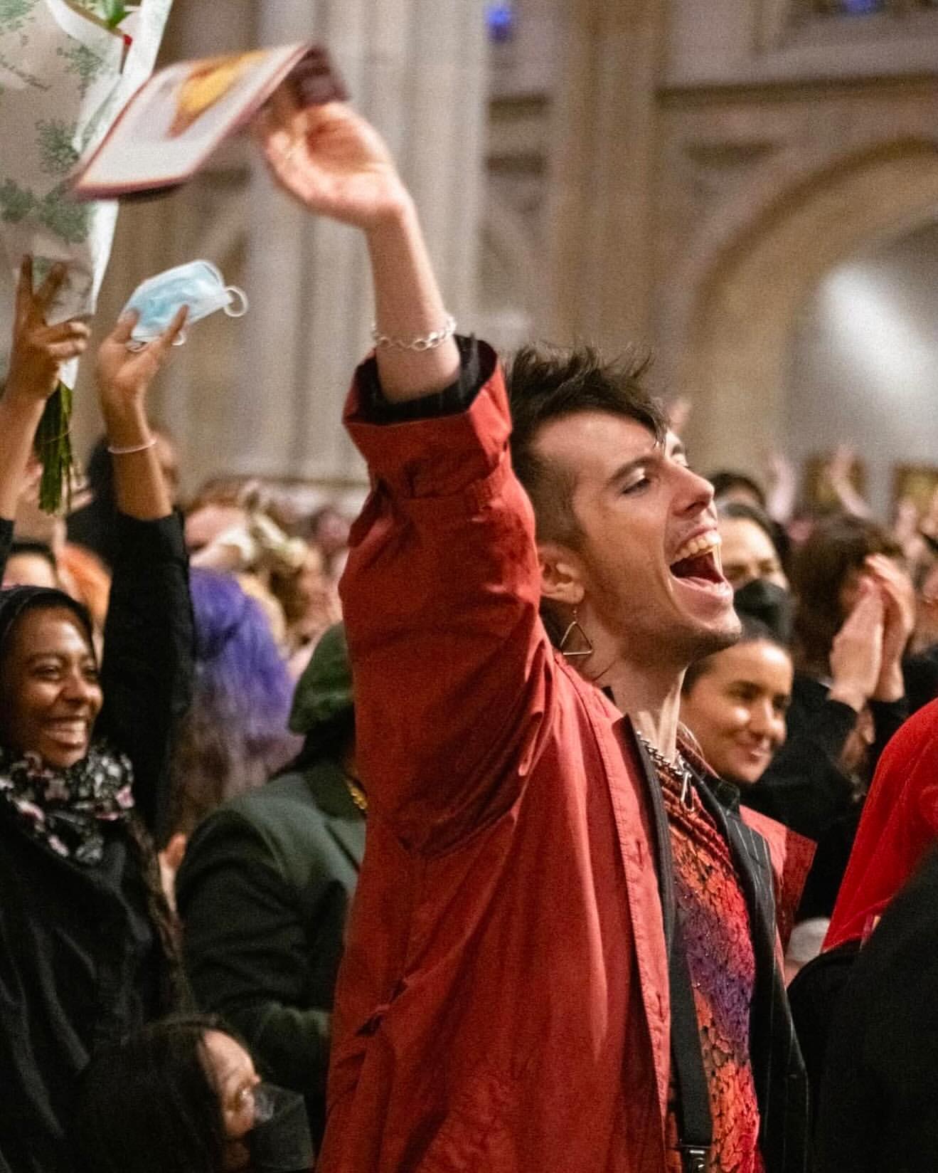 A diverse group of people celebrates indoors, raising hands and objects like a face mask. The setting appears to be a church interior.