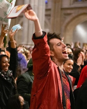 A diverse group of people celebrates indoors, raising hands and objects like a face mask. The setting appears to be a church interior.