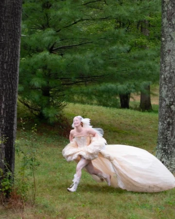 A person in a voluminous light-colored gown running through a green woodland setting framed by large trees.