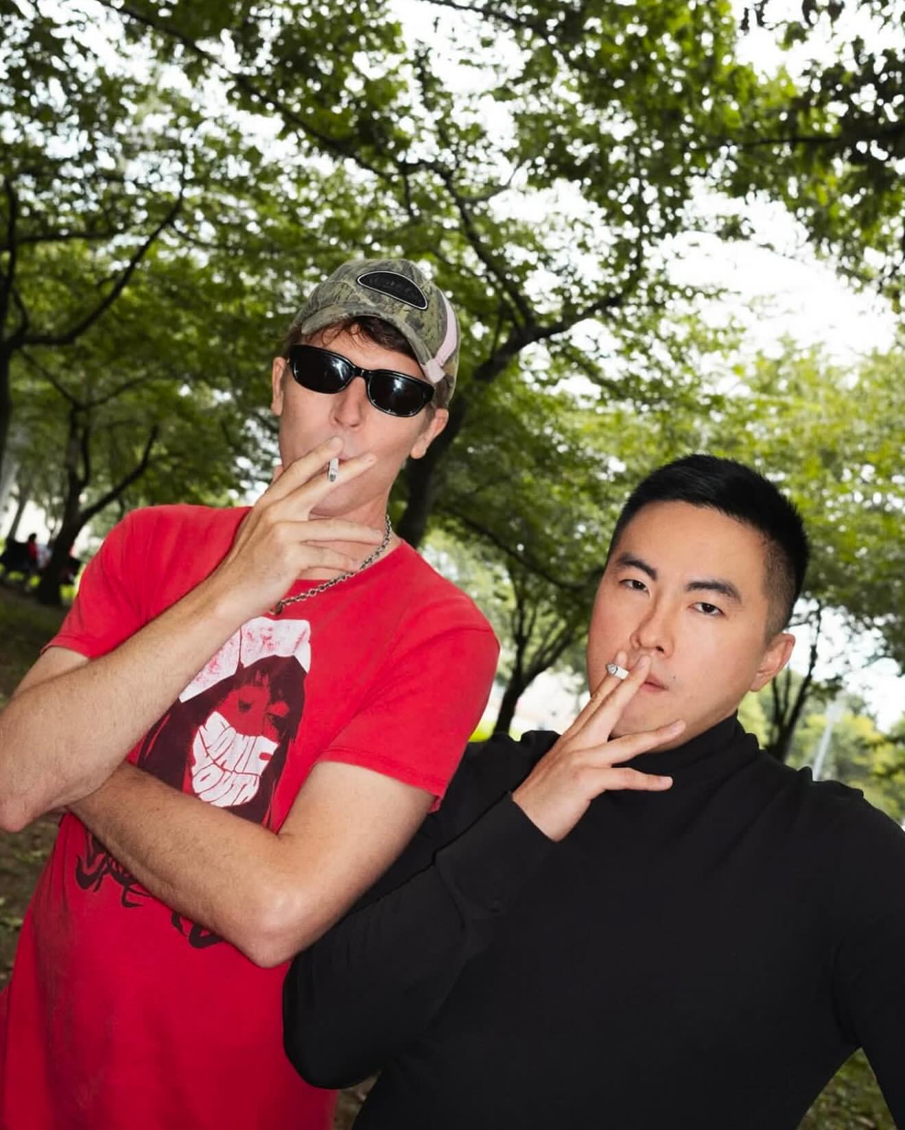 Two men are smoking cigarettes in an outdoor environment surrounded by green trees. One man is wearing a camo hat and sunglasses.