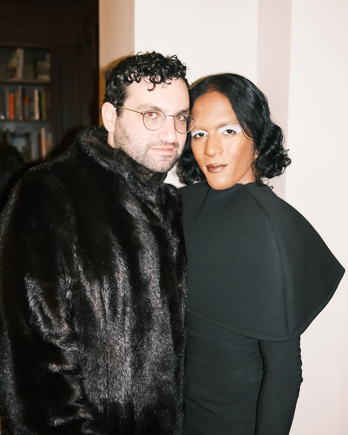 A stylish couple poses indoors; one in a fur coat and glasses, the other in a black dress with striking white makeup.