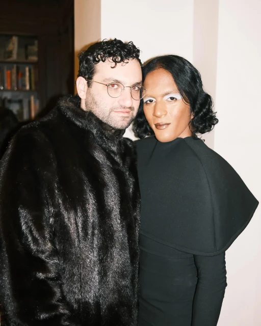 A stylish couple poses indoors; one in a fur coat and glasses, the other in a black dress with striking white makeup.