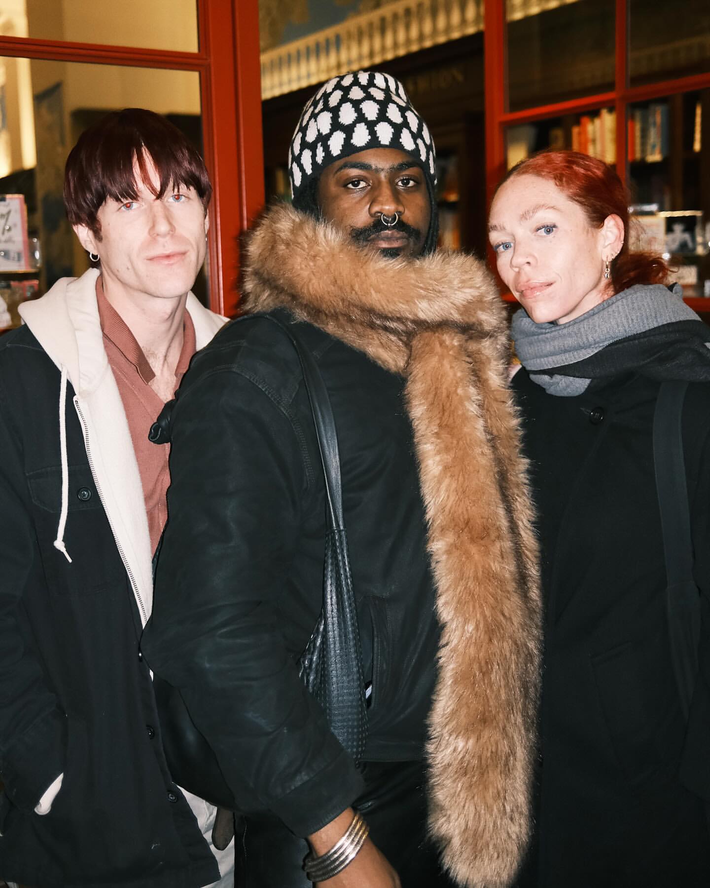 Three people stand together indoors, dressed in winter clothing. Two are wearing jackets, and the person in the middle has a fur stole.