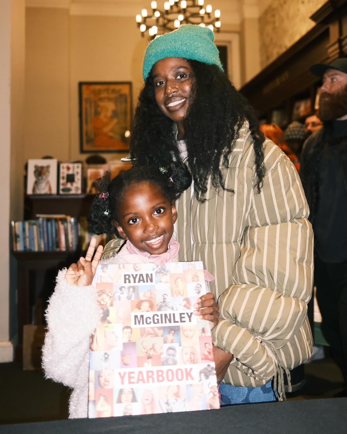 A young girl and adult woman pose with a book titled Ryan McGinley Yearbook, inside with warm indoor lighting and visible bookstore background.