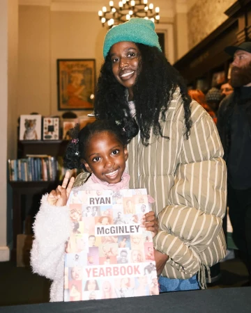 A young girl and adult woman pose with a book titled Ryan McGinley Yearbook, inside with warm indoor lighting and visible bookstore background.
