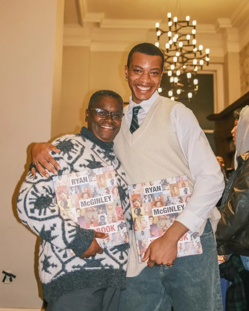 Two smiling people pose indoors holding Ryan McGinley books, showcasing fashion and art. Chandelier visible in background.
