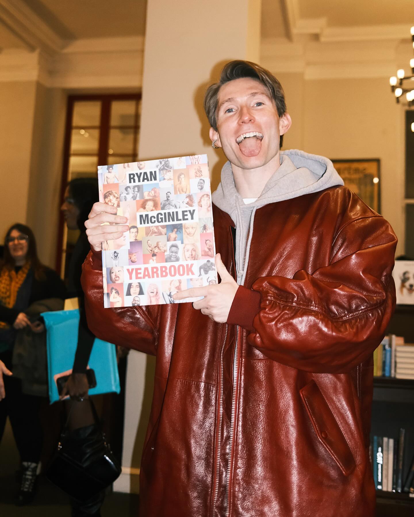 Man in brown jacket holding a book titled Ryan McGinley Yearbook in an indoor setting with another person in the background.