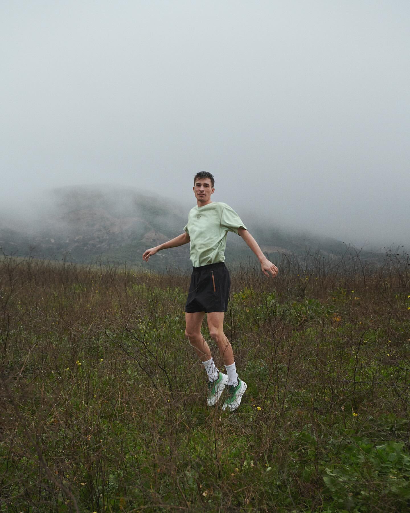 Man in athletic wear jumps in a grassy field with a mountain shrouded in fog in the background. Cloudy, overcast day.