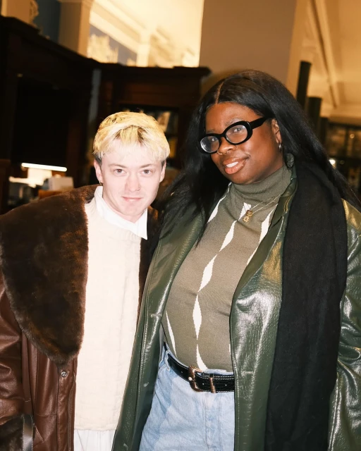 Two adults pose indoors. One with blond hair and a brown coat and one with long black hair, glasses, and a green coat.
