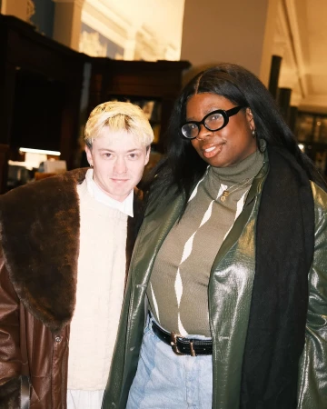 Two adults pose indoors. One with blond hair and a brown coat and one with long black hair, glasses, and a green coat.