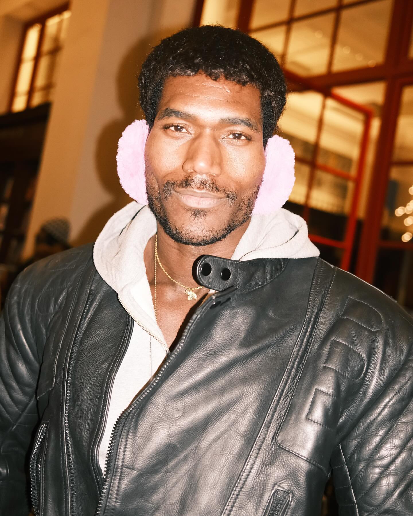 Adult Black man wearing pink earmuffs, leather jacket, and gold chain smiles in an indoor environment with a window background.