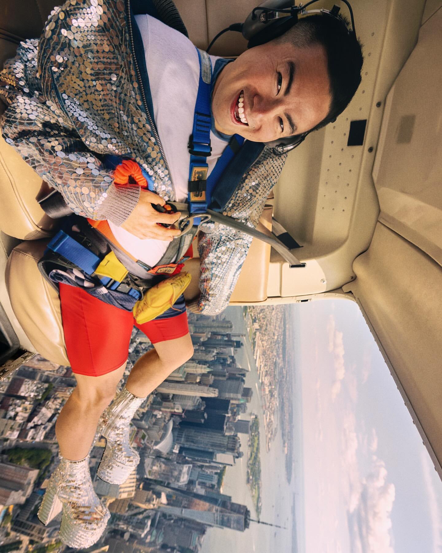 Smiling man in sparkling jacket inside helicopter with headphones, red shorts, metallic boots, cityscape backdrop, adventure travel vibes.