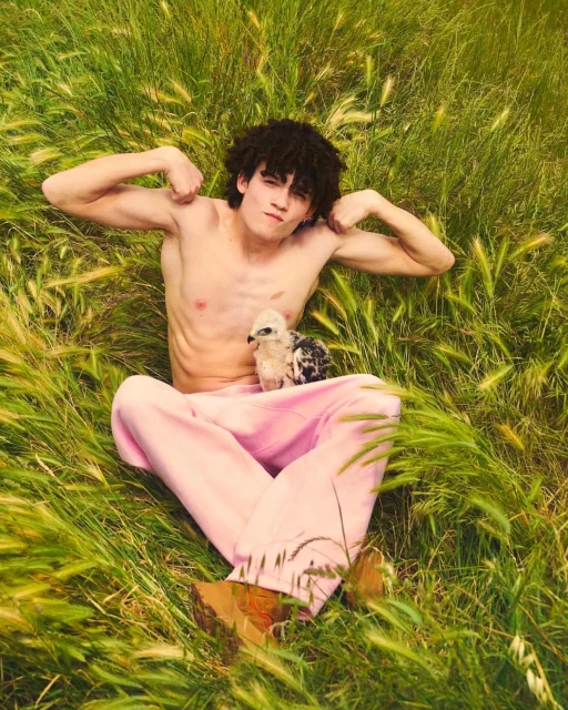 An adult man poses in a grassy field with an owl chick perched on his legs. He is shirtless, wearing pink pants, and flexing his arms.