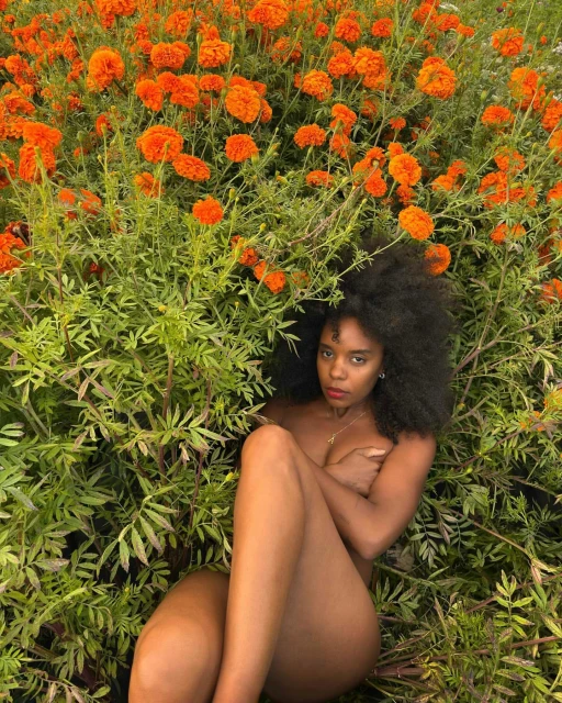 A nude woman with afro hair rests in a bed of bright orange marigold flowers, with a serious expression, and tan skin in natural light.