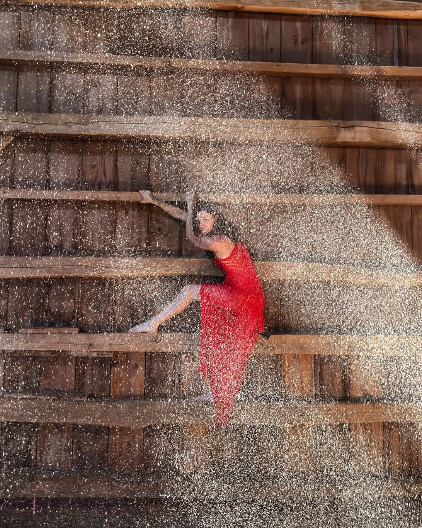 Woman in red dress strikes a dramatic pose on weathered wood stairs. Glittering particles create a visually arresting, textured effect.