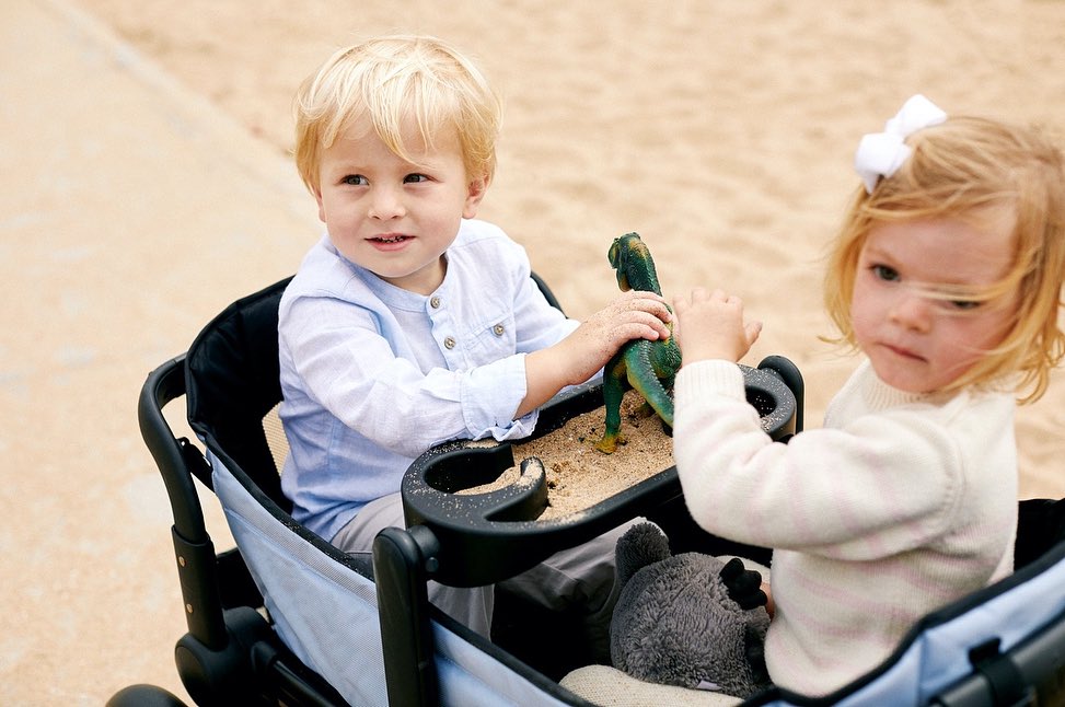 Two young children, a boy and a girl, sit in a wagon, playing with a toy dinosaur and stuffed animal on a sandy surface.