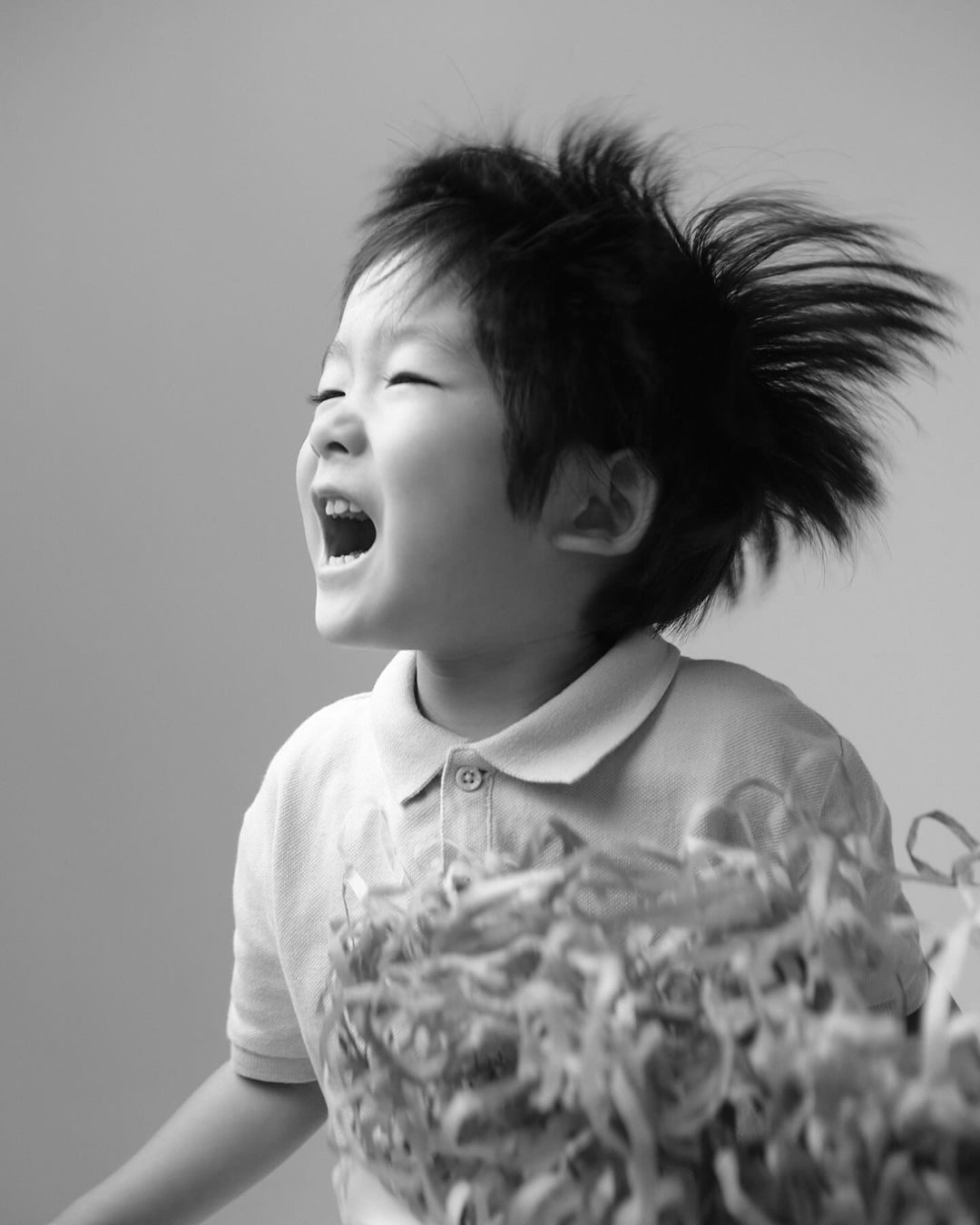 A child in motion, captured in black and white, showcases energy as his hair flies and he smiles widely, set against a plain background.