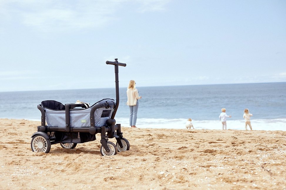 Beach day featuring a grey stroller wagon with an adjustable handle and a small group of people in the background against a backdrop of blue sea.