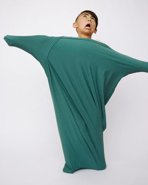 A young person with open mouth and outstretched arms is inside of a large green cocoon of fabric on a white backdrop in a studio setting.
