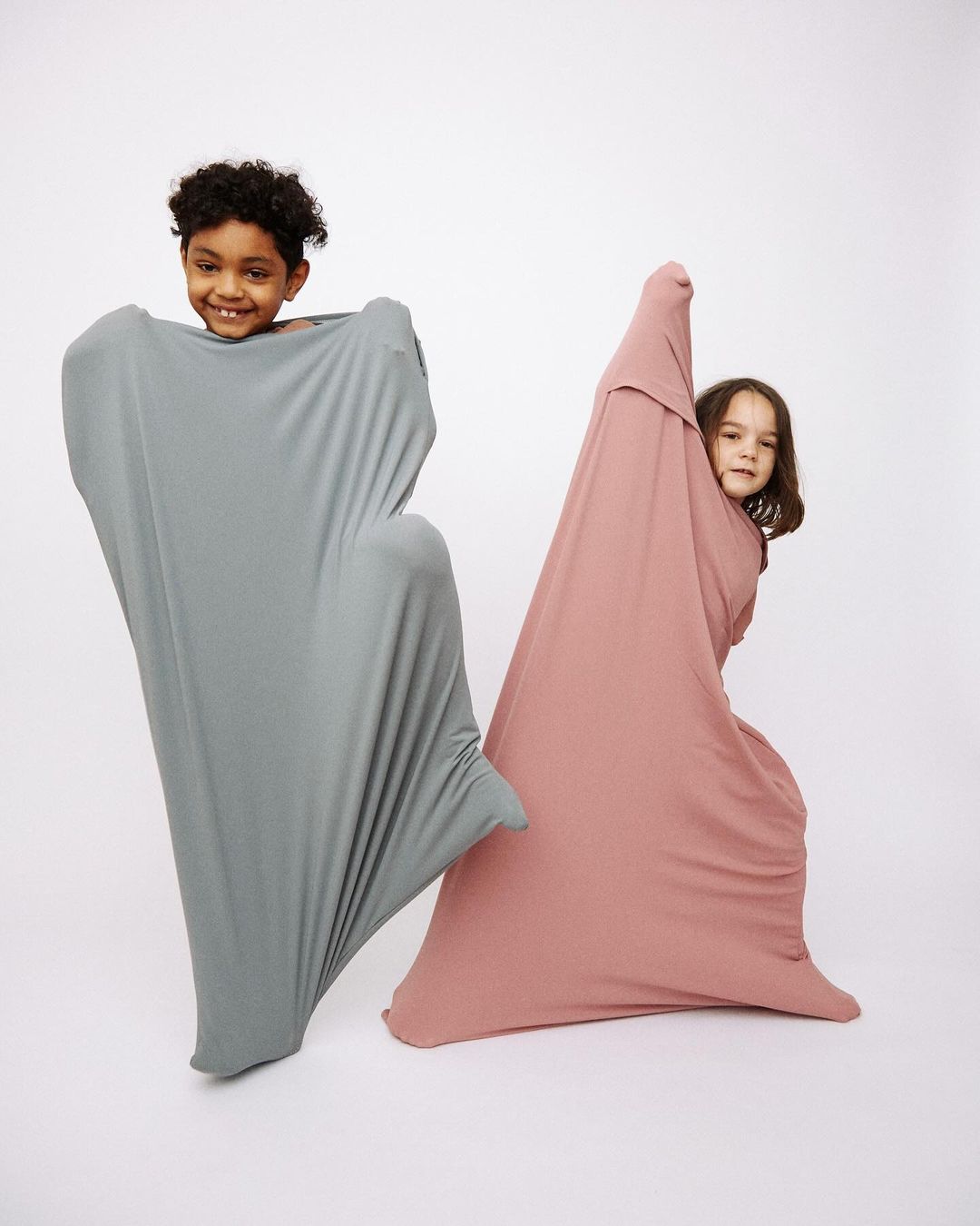 Two happy children are playing inside stretched fabric tubes in a light studio, creating playful shapes against a white background.