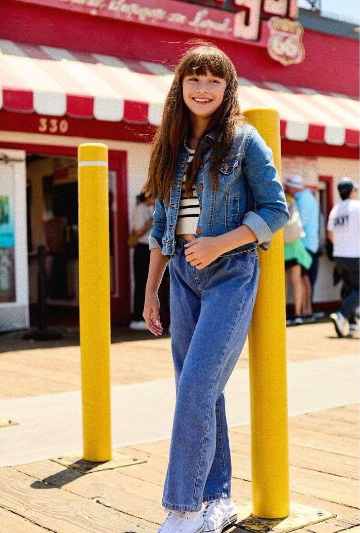 A young teenage girl stands in front of a building in a denim jacket and jeans with an upbeat expression and brown hair.