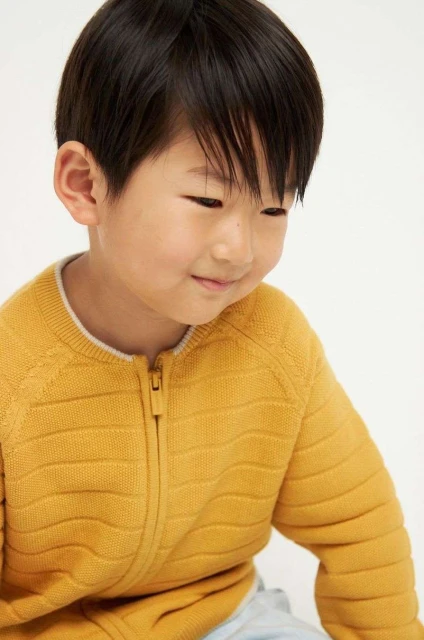 Close-up portrait of an Asian child in a yellow zip-up sweater, smiling slightly, against a plain white background in a studio setting.