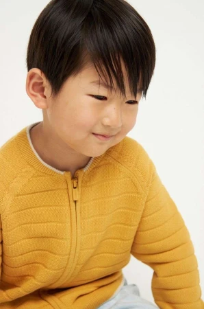 Close-up portrait of an Asian child in a yellow zip-up sweater, smiling slightly, against a plain white background in a studio setting.
