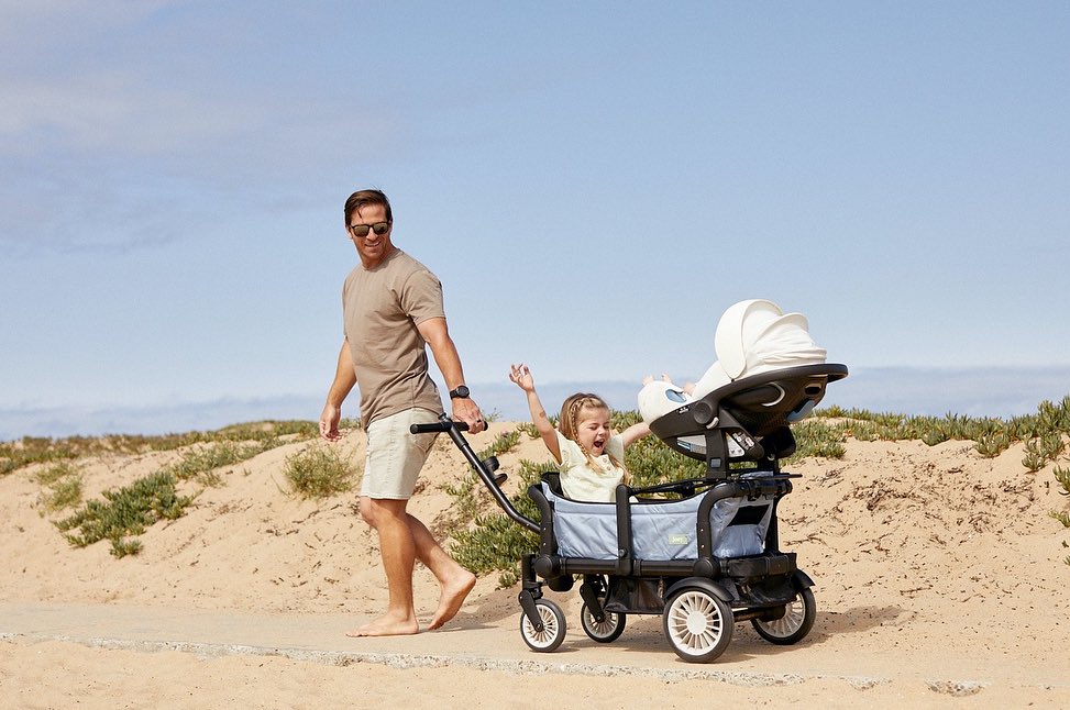 Adult male pulls a wagon with a young girl and a covered infant car seat on a sandy path with beach vegetation on a sunny day.