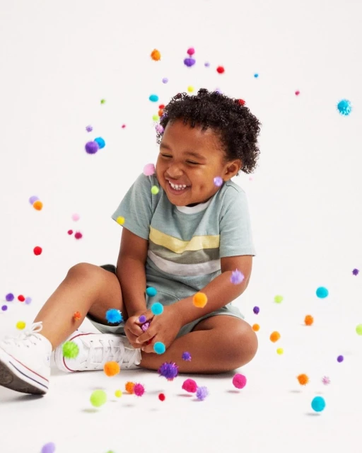 Young child seated indoors, smiling and surrounded by colorful pom poms in mid-air against a white background, creating a playful scene.