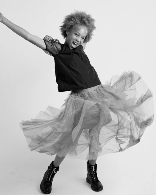 Young girl with curly hair in black top, layered tulle skirt, and boots, striking joyful pose in bright studio. Fashionable and fun.