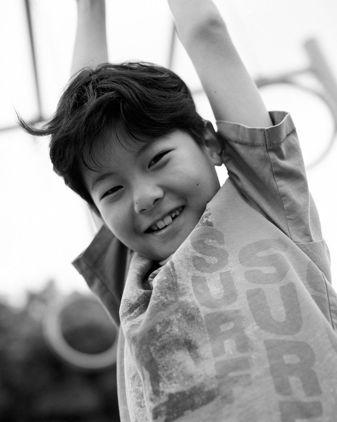 Monochrome portrait of a young boy hanging from a playground structure, smiling with an open mouth. The image is bright with soft focus.