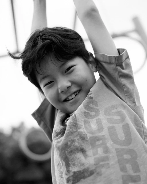 Monochrome portrait of a young boy hanging from a playground structure, smiling with an open mouth. The image is bright with soft focus.
