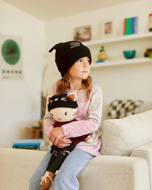 Young girl with long hair sits on a sofa holding a doll dressed in a matching knit hat indoors with natural light.
