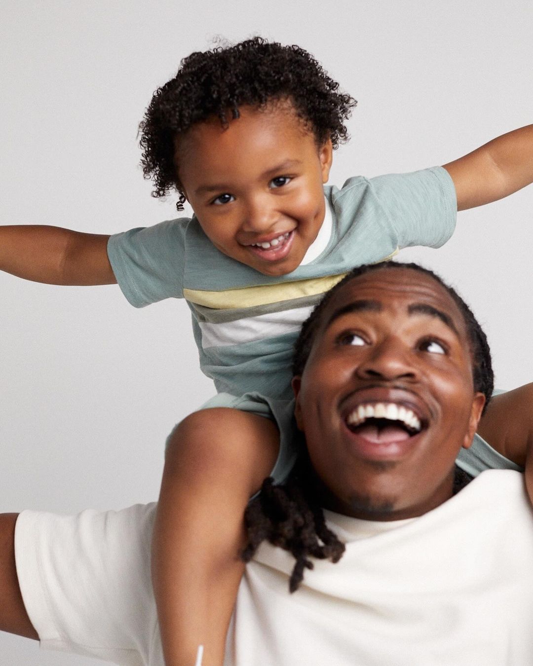 Smiling Black father and son in matching t-shirts. Boy sits on his dad's shoulders, arms outstretched against white backdrop.