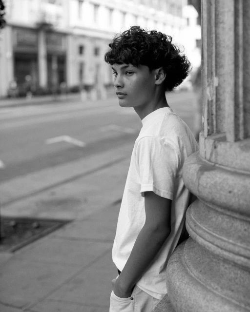 Monochrome portrait of a young adult resting against a column in an urban setting, looking off to the side with a pensive expression.