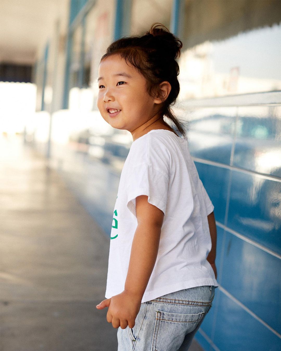 Smiling Asian child standing in a hallway wearing a white t-shirt and denim jeans, looking off to the left, bathed in natural light.
